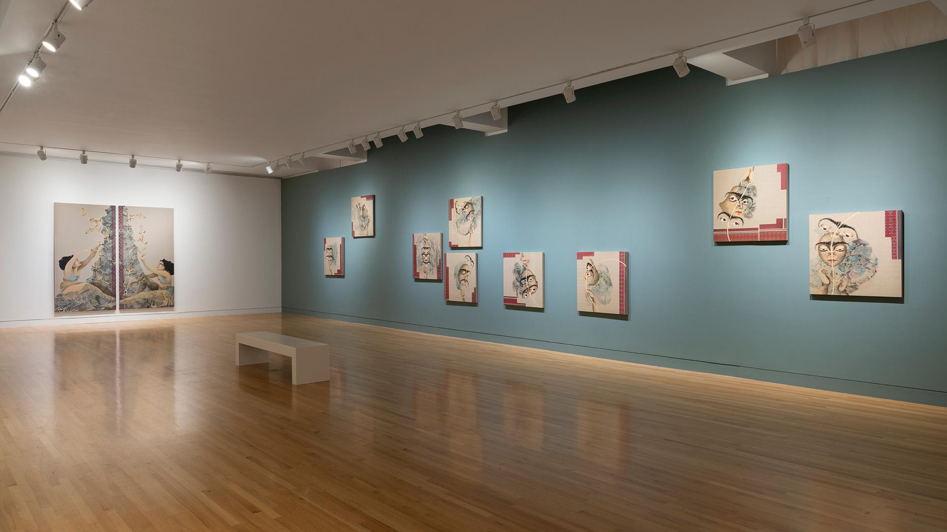 Two walls in a large gallery, one painted blue and covered in a seemingly haphazard manner with square paintings from a series focused on eyes and faces, the other white with a large artwork made from two panels