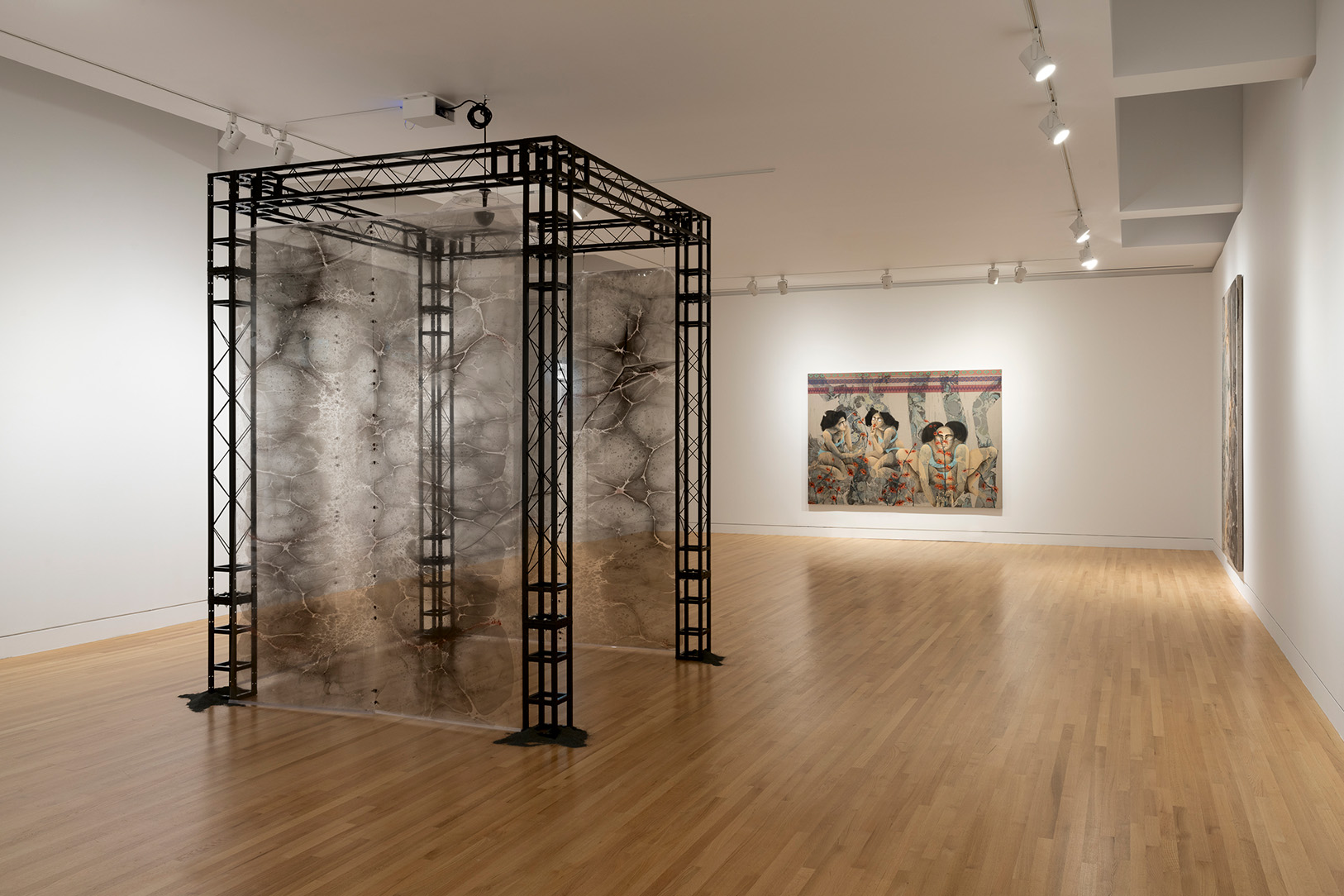 A gallery space containing a rectangular, freestanding structure large enough for a person to walk into with a metal frame and sheer curtain walls, plus different artworks hanging on walls in the background