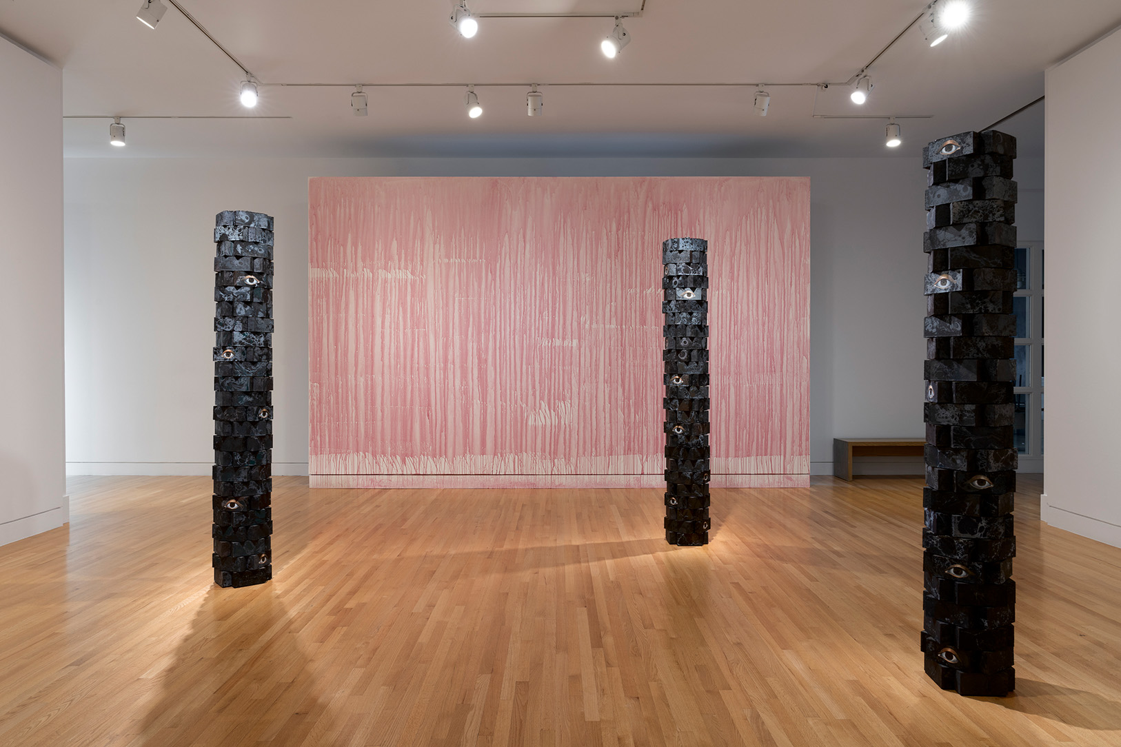 Three pillars made of stacked bricks painted a marbled blue with eyes appear at irregular intervals in front of a lightly painted pink wall with visible drips of paint