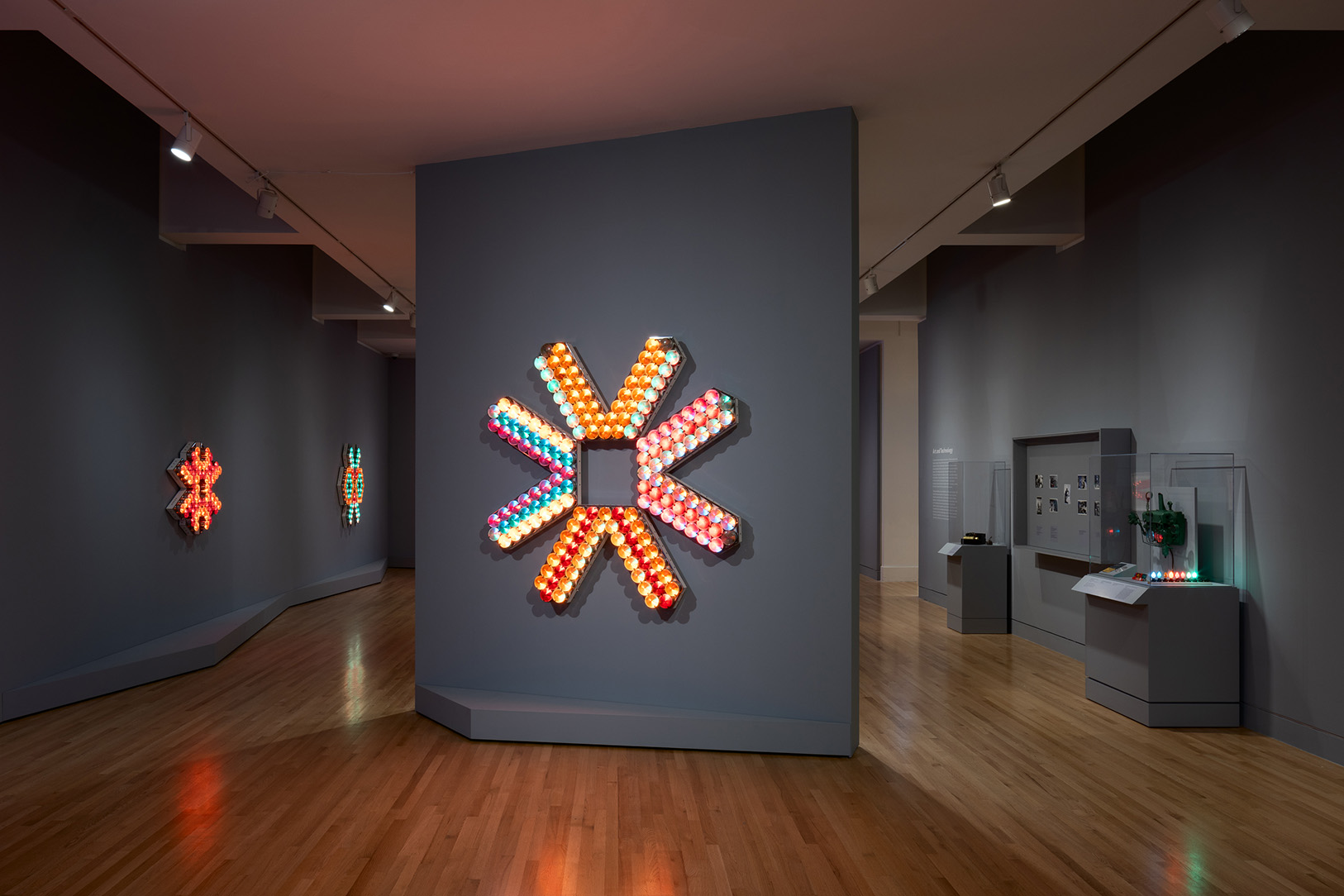 A large, geometric artwork made up of a number of individual light bulbs mounted on an angled wall in a gallery space.