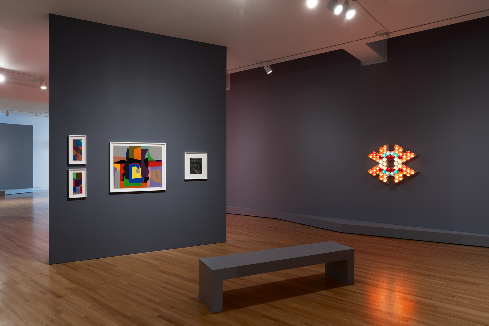 Artworks in a gallery space including a geometric artwork made up of a number of individual light bulbs and flat, abstract works on paper.
