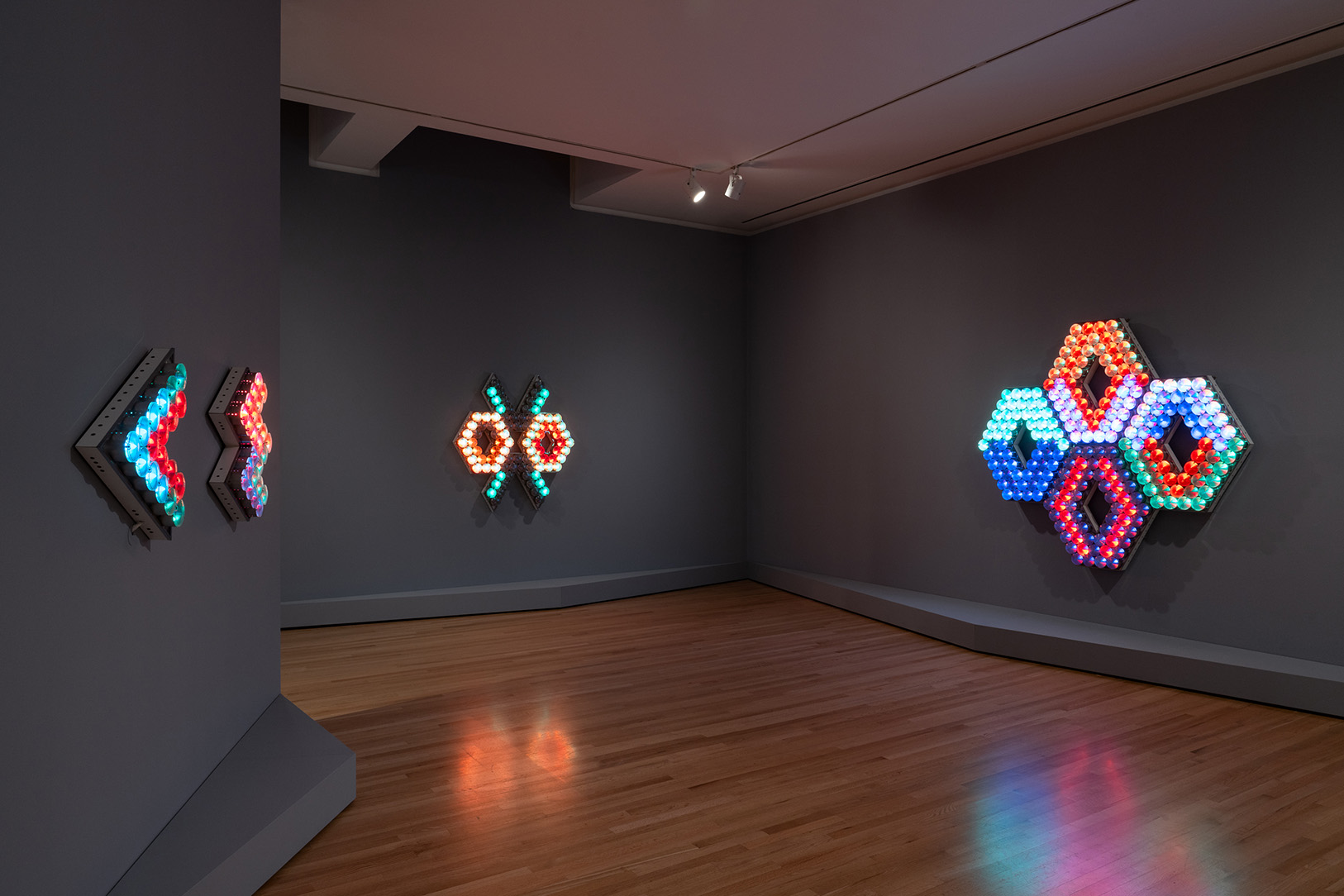 Three large, geometric artworks made up of a number of individual light bulbs mounted on an angled wall in a gallery space.
