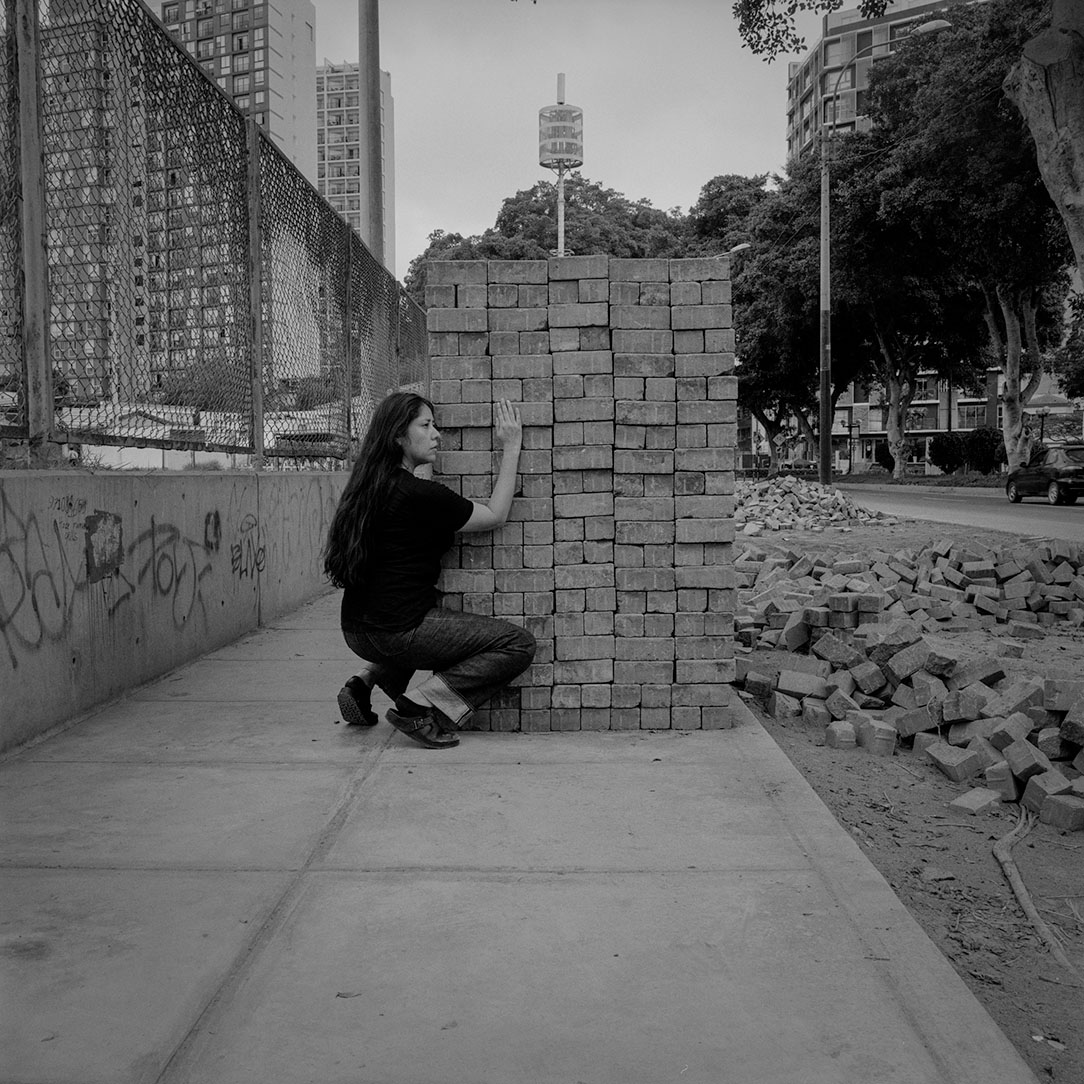 A black and white film photograph of a woman croching and posting in front of a pile of bricks in an urban area