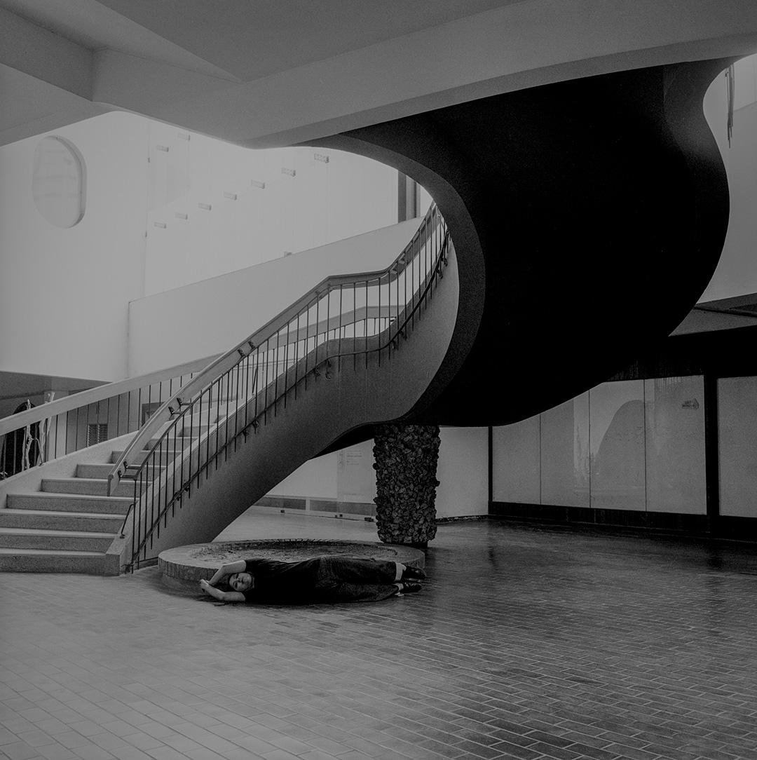 A black and white film photograph of a woman lying on her side facing away from the viewer in front of a large circular staircase