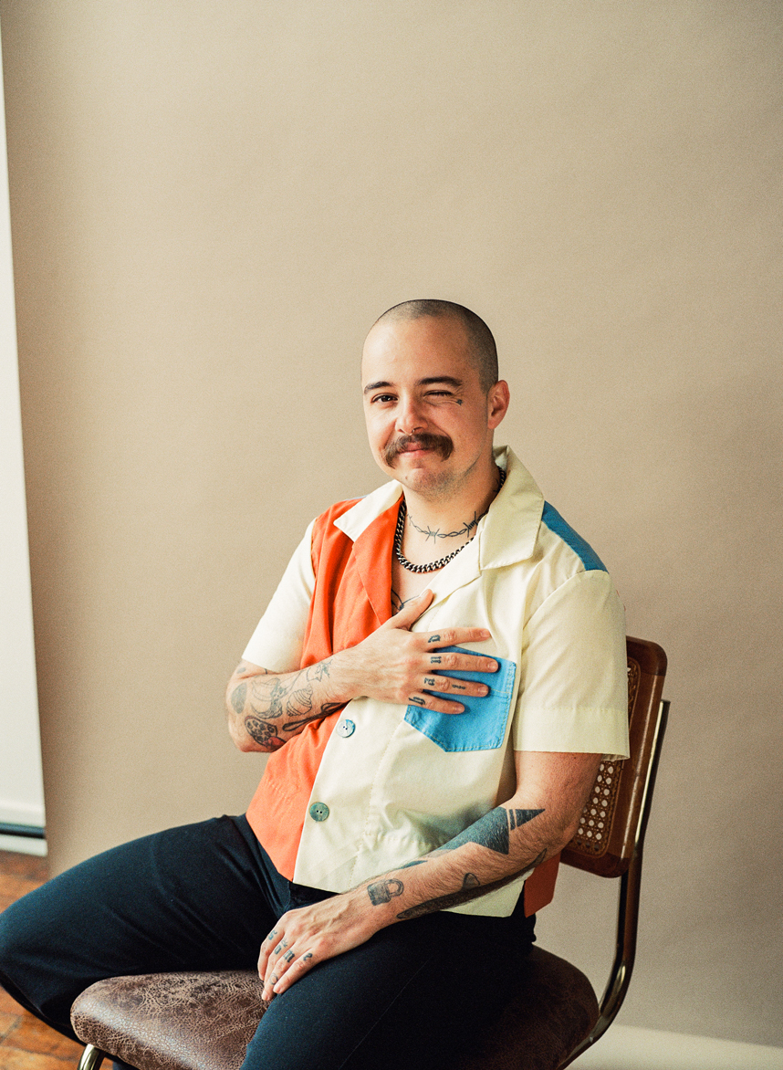 Lane Bestold sitting in chair wearing a colorful button up shirt winking and holding his hand over his heart
