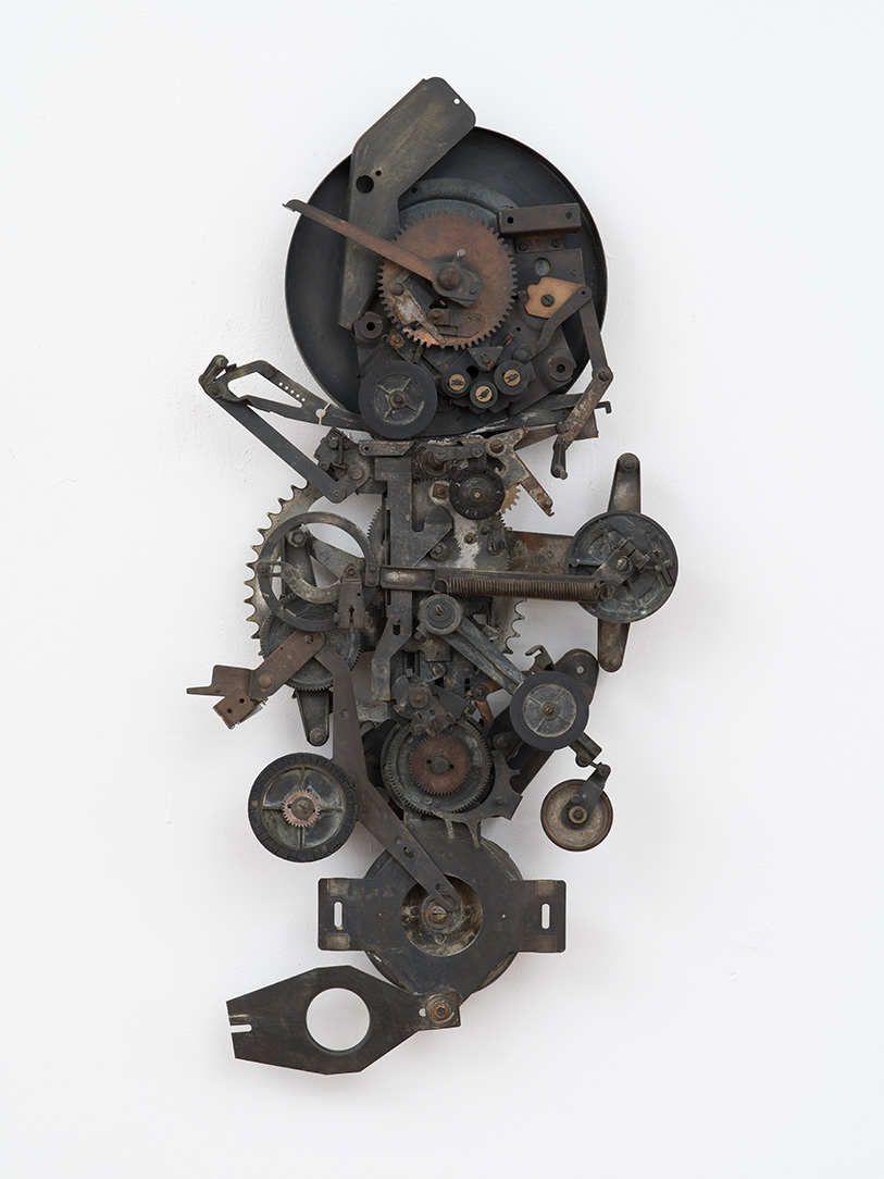 A artwork on a wall made up of darkly colored metal mechanical gears and objects