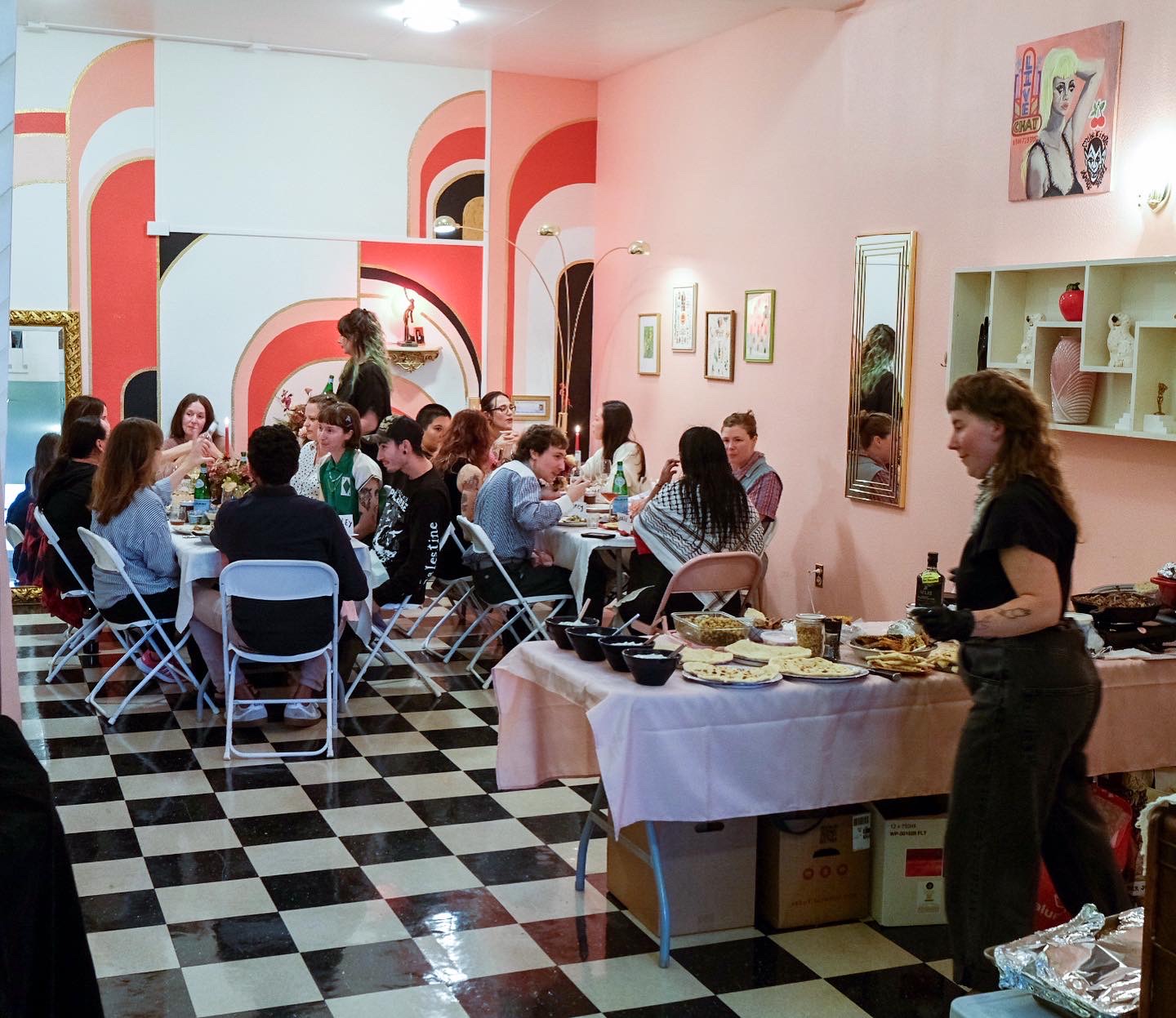 People sitting at two tables. In the foreground a table is laid with food and Cece Edson is doing prep work