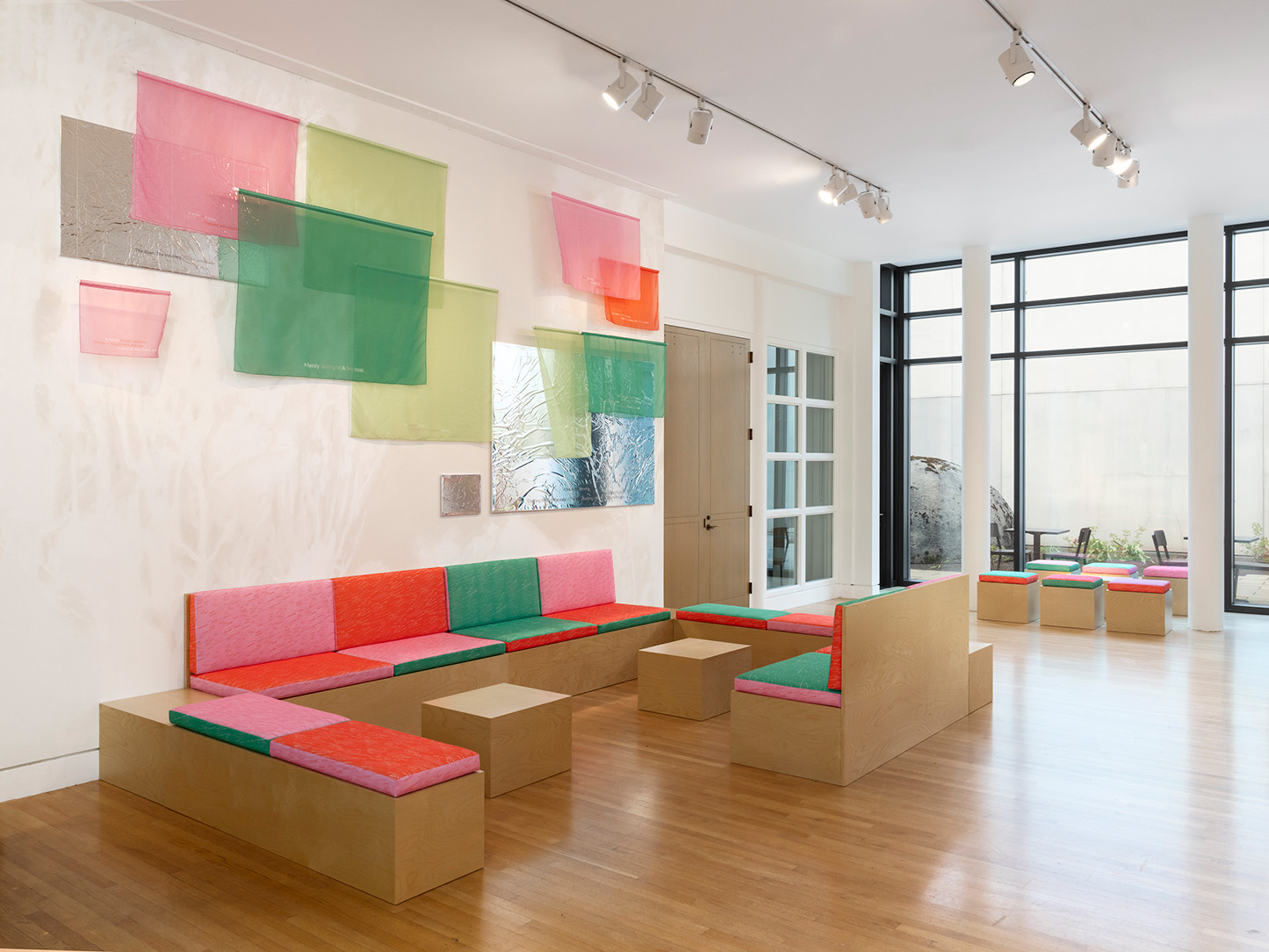 A large interior space, well let through a large glass wall, with low benches and colored fabric patterns on the walls