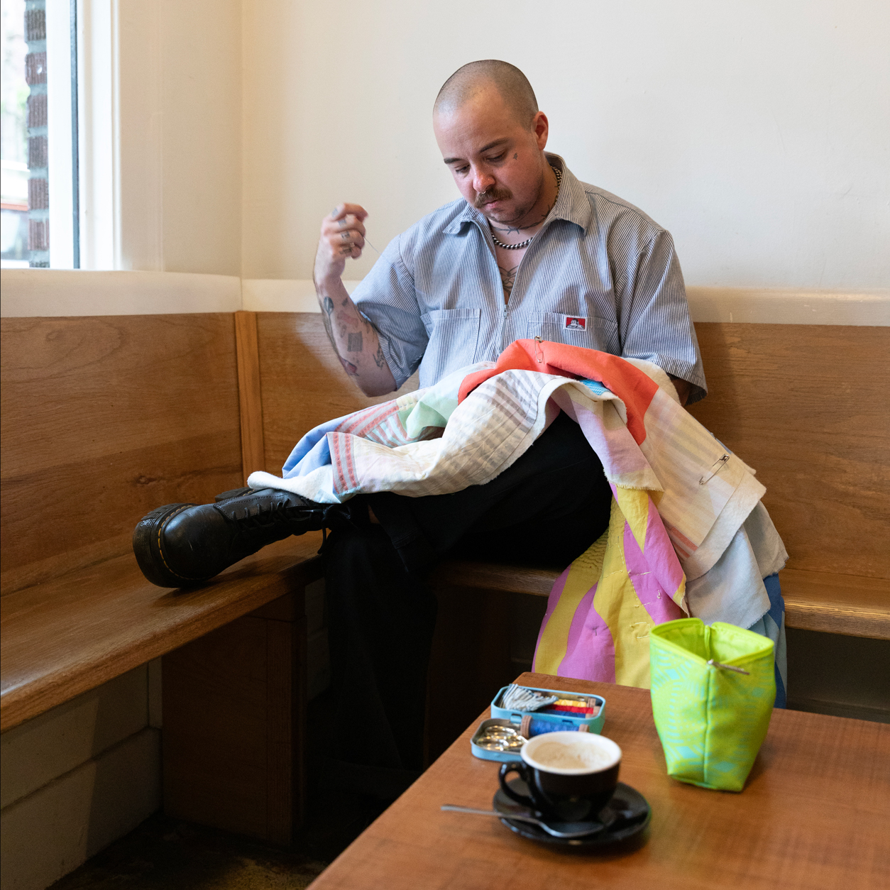 Lane Bestold handsewing a quilt on his lap while sitting in a corner booth