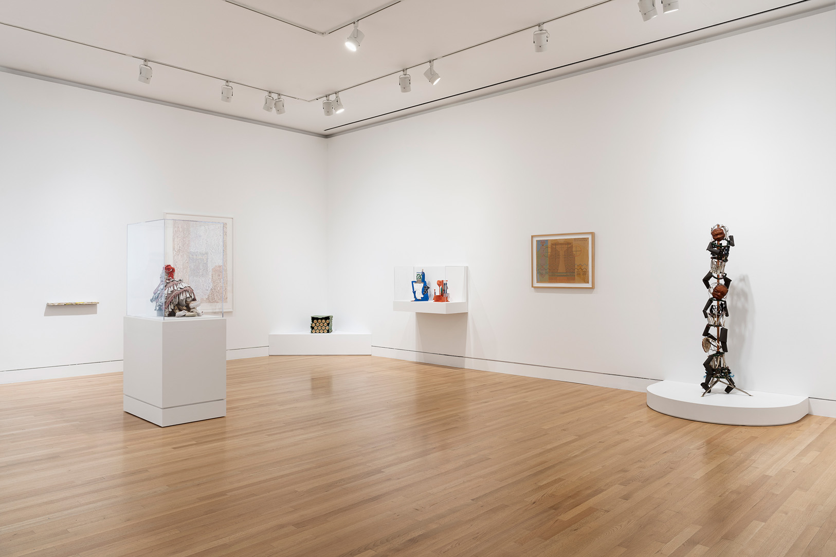 A view towards a corner of a gallery space with a variety of artworks, some on pedestals and shelves, and others hung on the walls
