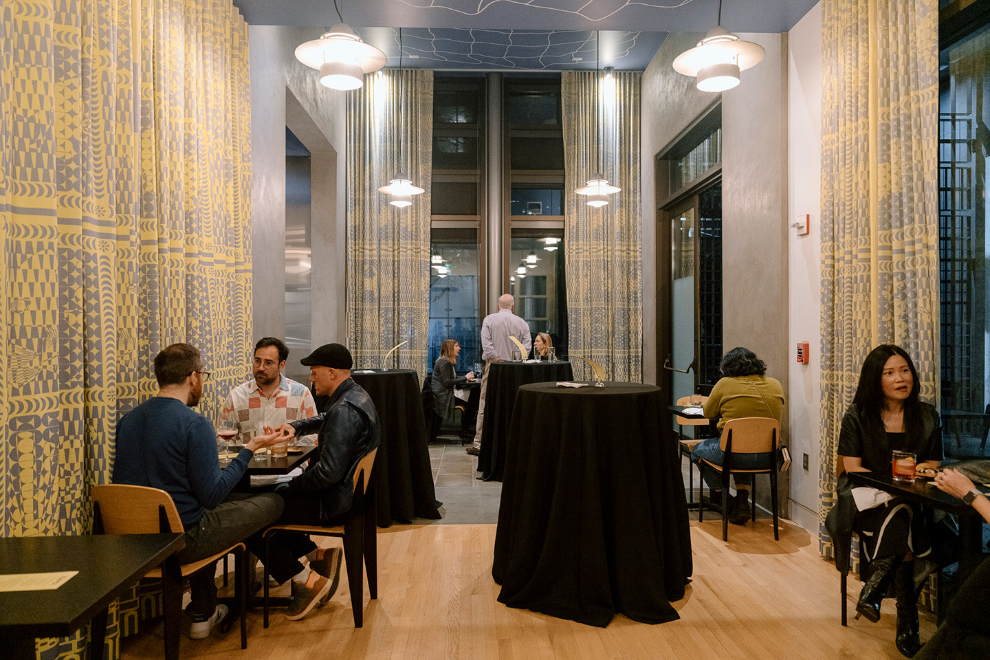 Small groups of people seated at tables in a café space hung with yellow curtains