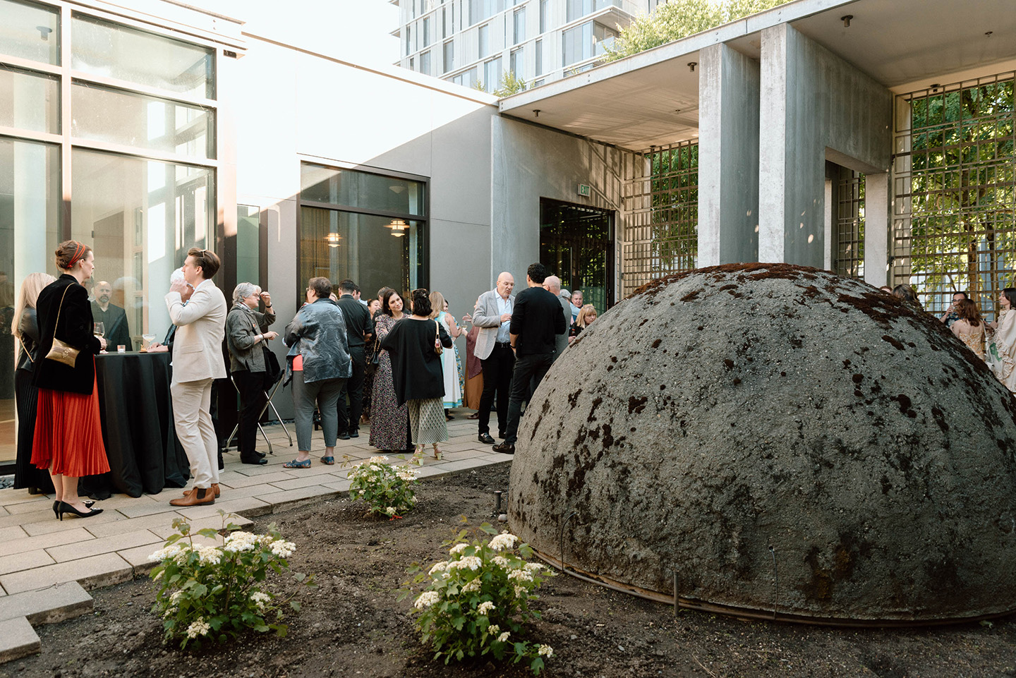 Groups of standing and chatting people in an open, outdoor courtyard on a sunny evening.