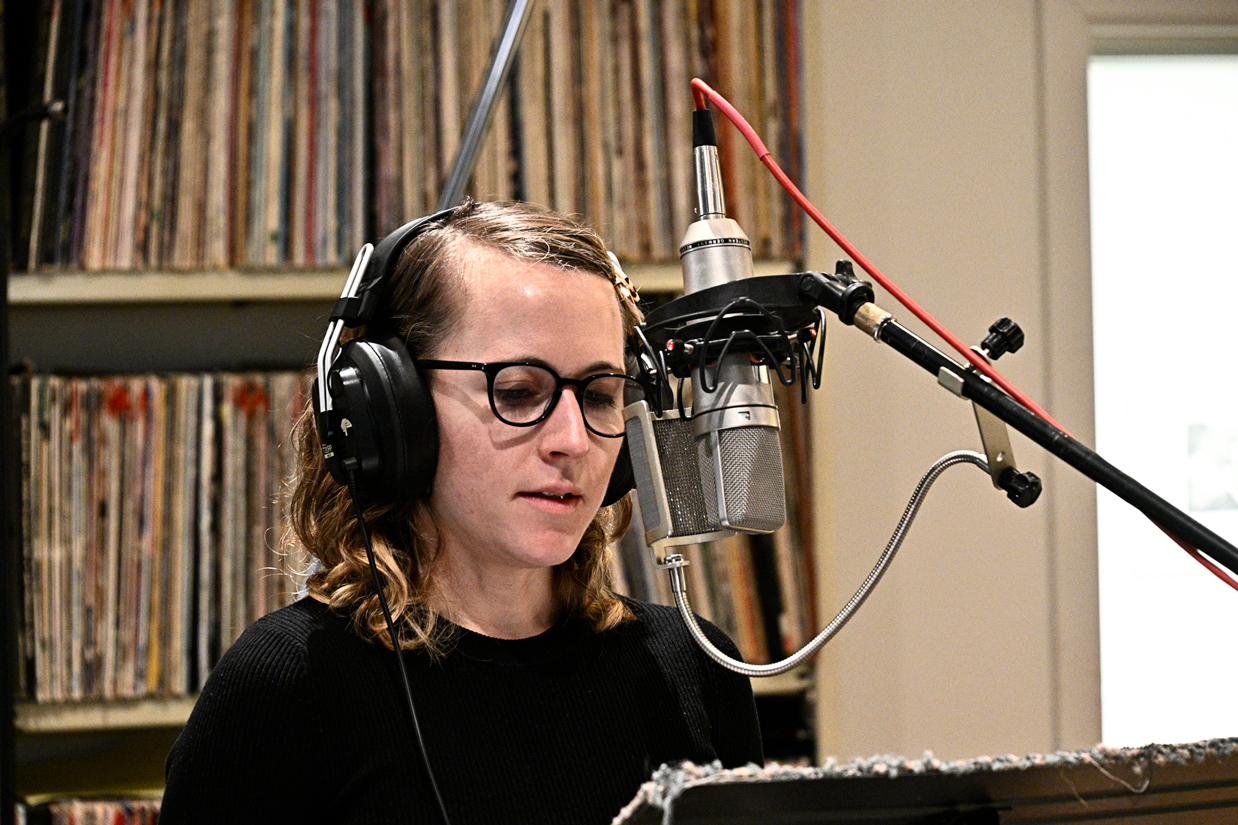 Photo of Erin Langner, a woman with shoulder length brown hair and glasses, smiling in front of a microphone in a recording studio