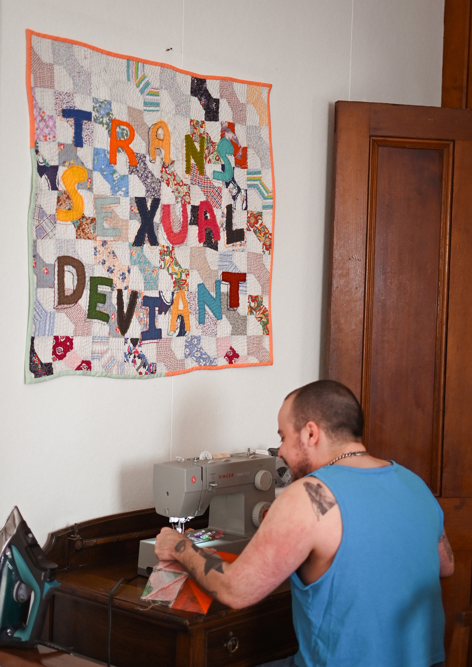 Lane Bestold sitting at a sewing machine sewing with a square quilt with the words "TRANS / SEXUAL / DEVIANT" hanging on the wall