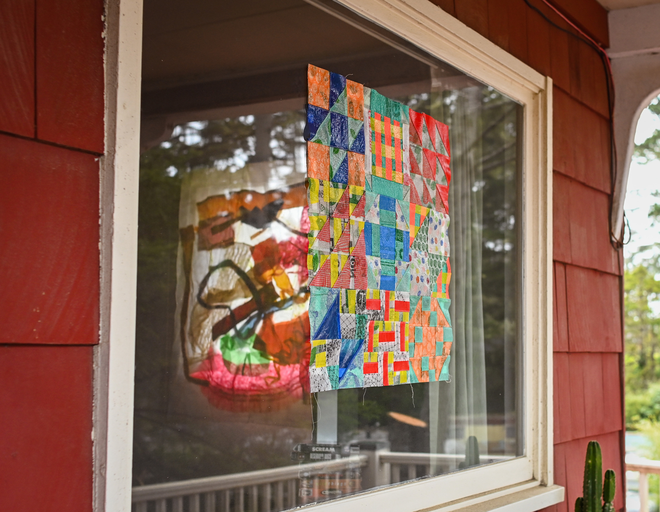 A colorful square quilt hanging in the window of a house. Another opaque quilt with organic shapes can be seen inside hanging in front of another window