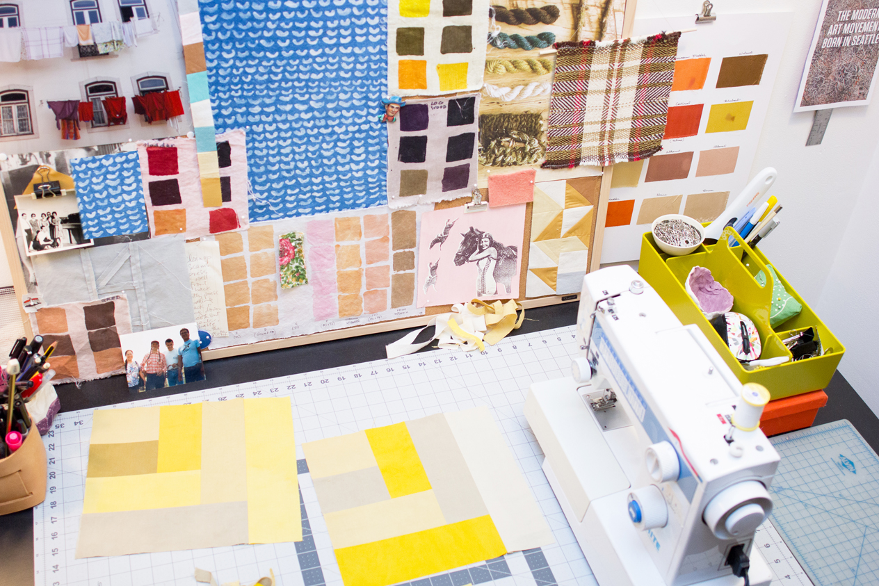 Photo of Naomi Parker's studio with a sewing machine and quilt squares on a table and a wall covered in swatches and bits of fabric