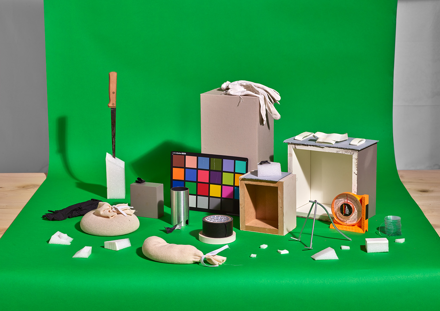 An assortment of objects including a color calibration chart, tape, boxes, styrafoam, and a knife on a chroma key green dropcloth
