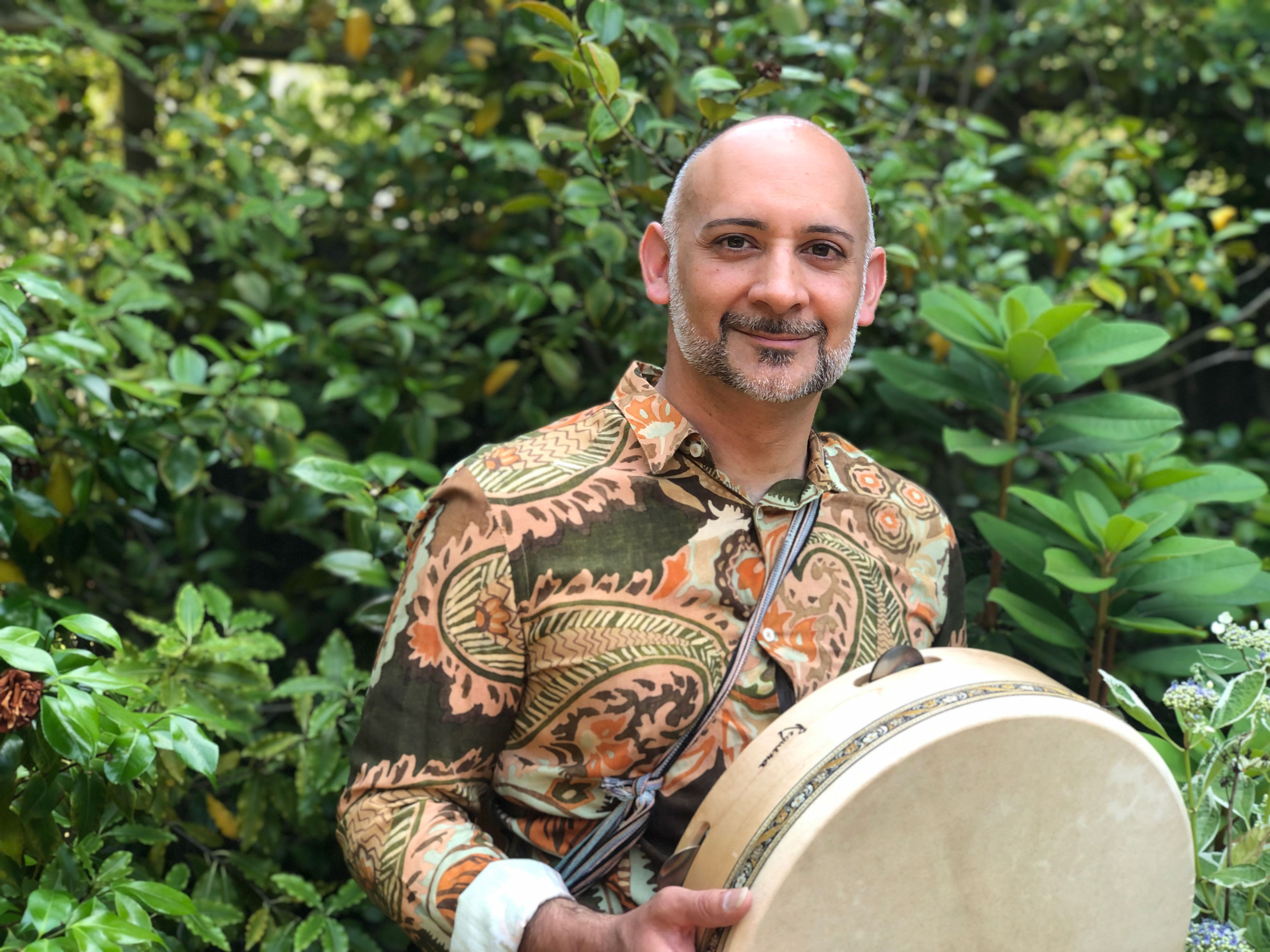Musician with a long sleeve patterned shirt holding a drum and standing in front of a tree