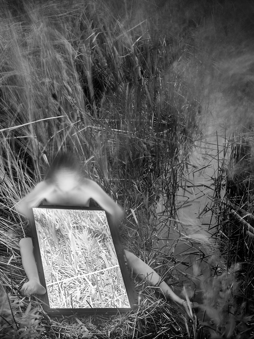 A black and white image of a nude human figure in a natural, reedy landscape with their body partially obscured by movement and a large LCD screen