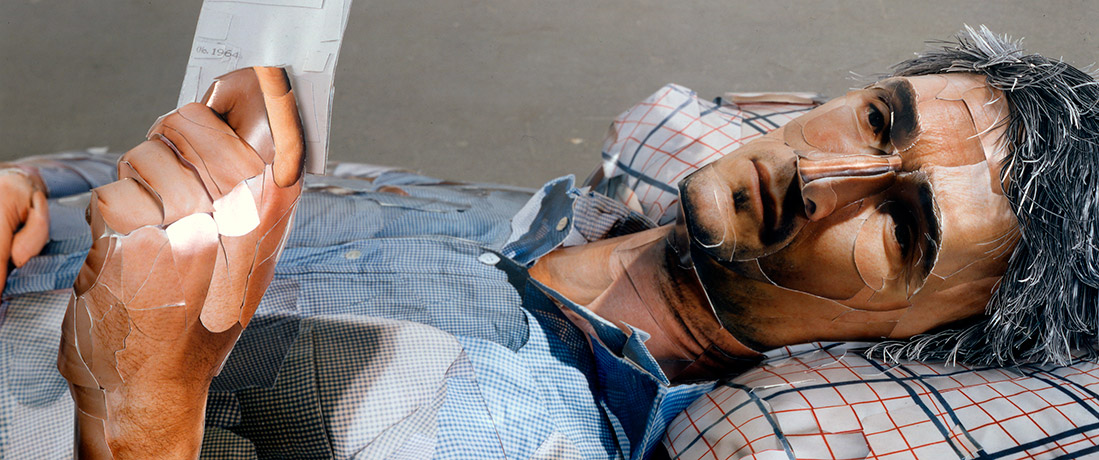 Detail of the face of a human figure made out of polystyrene and covered in prints to create the illusion of skin and clothing laying down with its knees up and reading from a piece of paper 