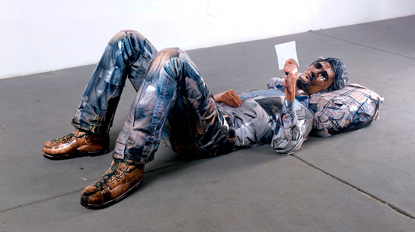 A human figure made out of polystyrene and covered in prints to create the illusion of skin and clothing laying down with its knees up and reading from a piece of paper 