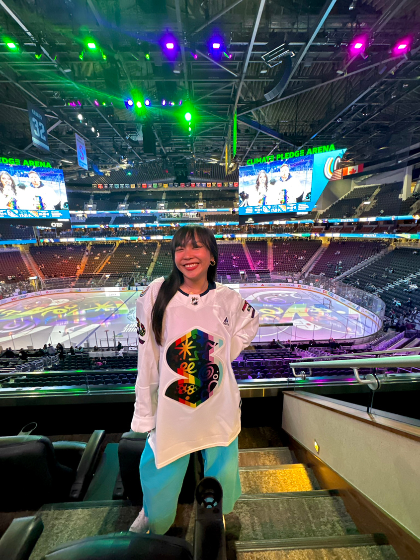 Photo of Nikita Ares, a woman with dark brown hair, wearing a Seattle Kraken jersey in front of the Kraken ice rink
