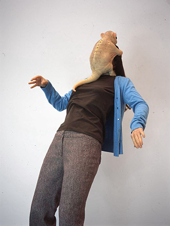 A realistic sculpture of a small, furless creature gripping a human figure's head and covering their face