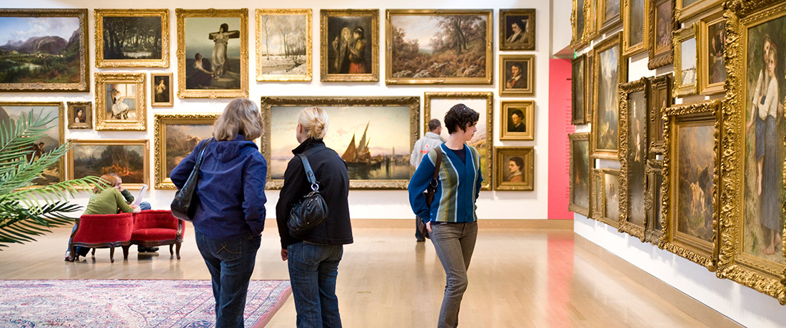 People in a large room hung with a carpet, chairs, and a potted plant with paintings hung floor to ceiling on the walls