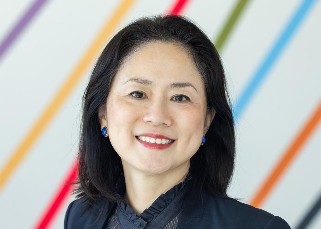 A professional headshot style photograph of a woman wearing formal clothing in front of a wall with colorful, diagonal stripes