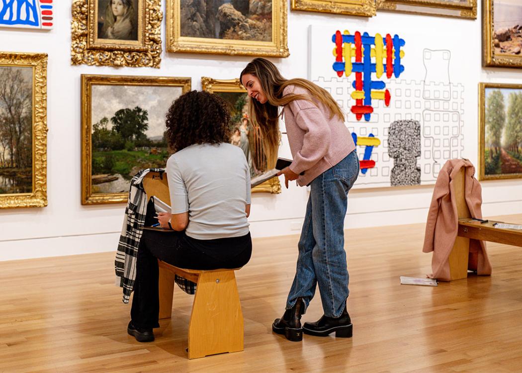 Two people chatting while holding clipboards in front of a large wall covered in paintings. One is sitting on a drawing stool and the other is standing nearby.
