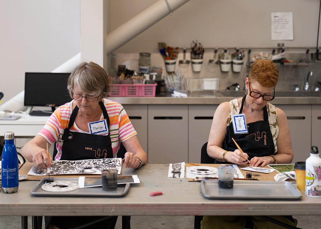 Two older people sitting side by side at a long table and working on art projects in a studio space