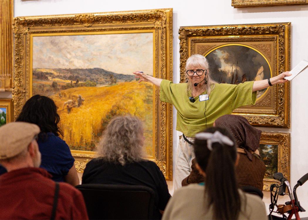 A person holding a notepad with their arms outstretched gesturing and presenting to a seated group of older adults in front of a wall covered in paintings