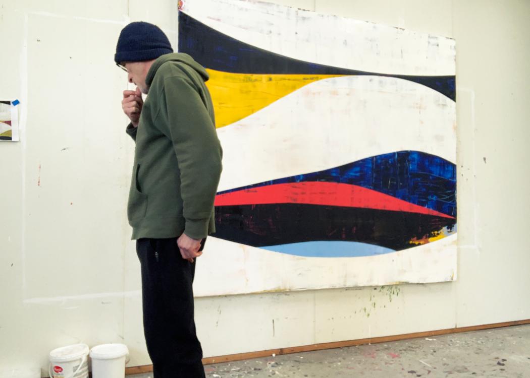 Photo of Ken Kelly, a man wearing a hoody and a beanie, working in his studio with one of his paintings on the wall behind him
