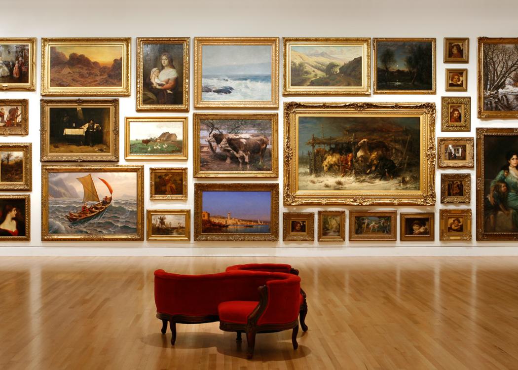 Museum gallery with paintings hung floor to ceiling and a red gossip chair on the floor in front