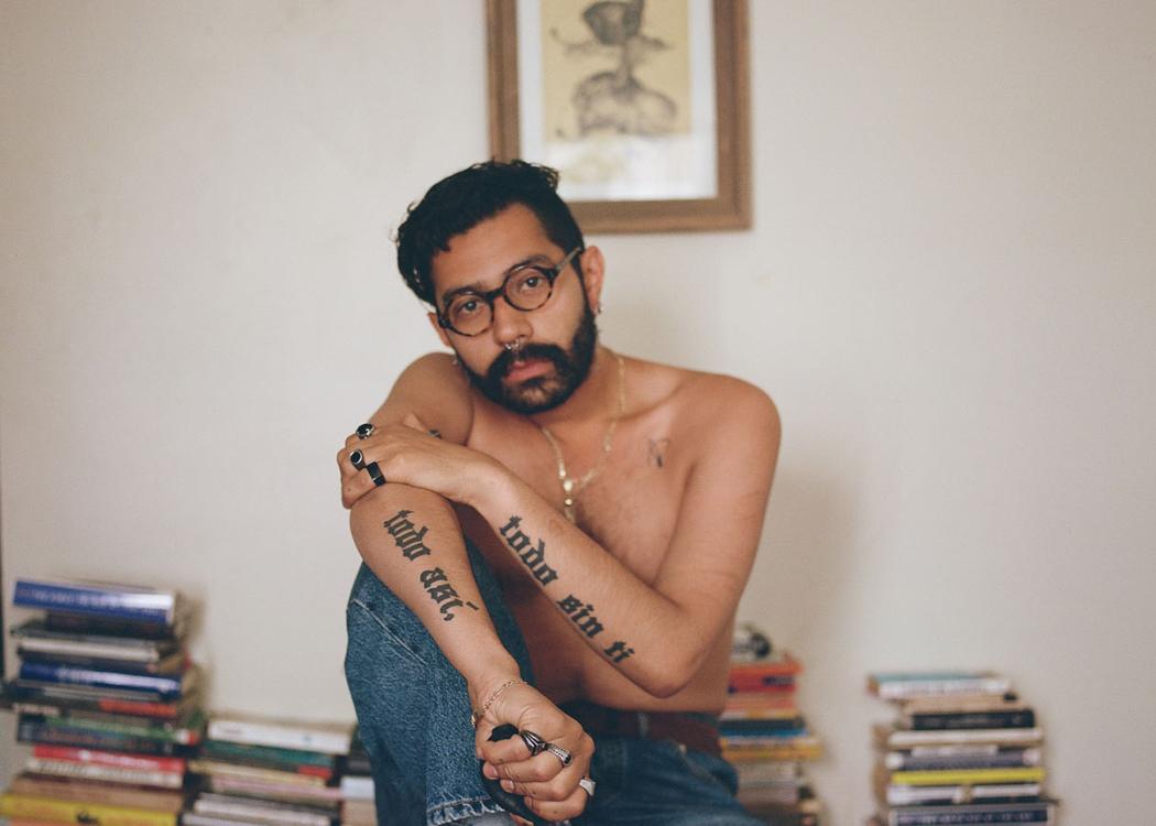 Portrait photo of a person wearing jeans and no shirt, sitting on a desk with books stacked behind them