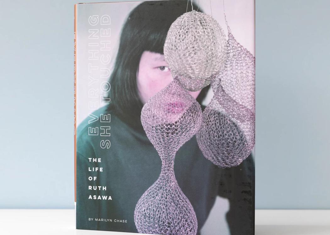 A book sitting on a white table, the cover features a woman's face slightly obscured by hanging metal artwork