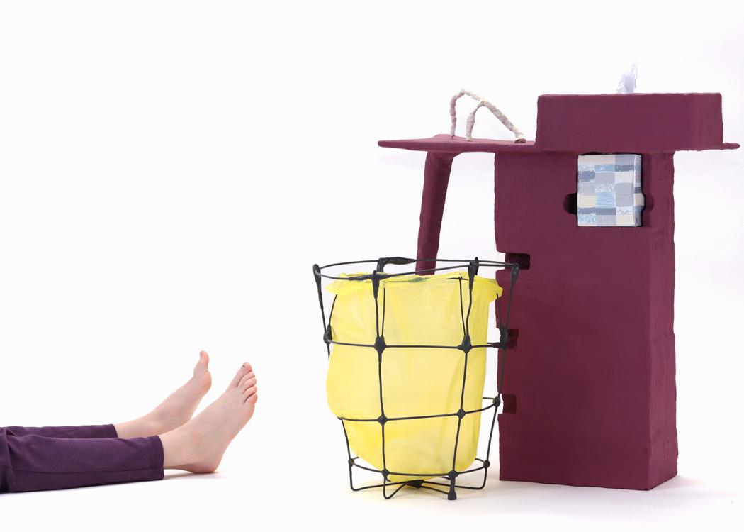 An artwork on the floor made up of a wire basket and a side table with a kleenex holder painted a deep maroonish purple. A small pair of legs and feet appear from the left of the image