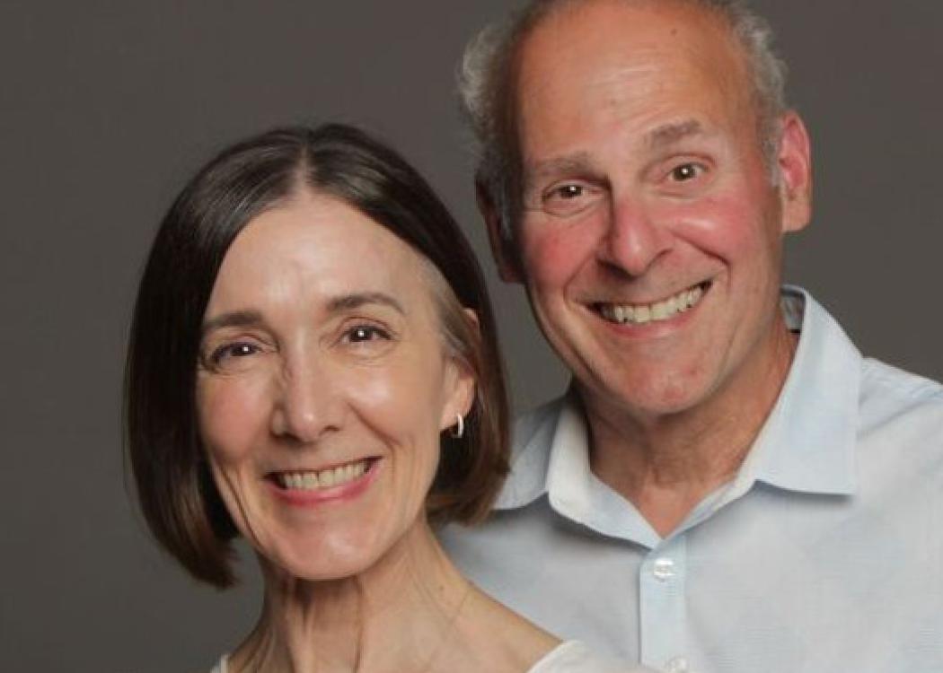 A professional headshot style photo of two people. In front is an older woman wearing a white shirt, and just behind and to the side is an older man wearing a light blue shirt.