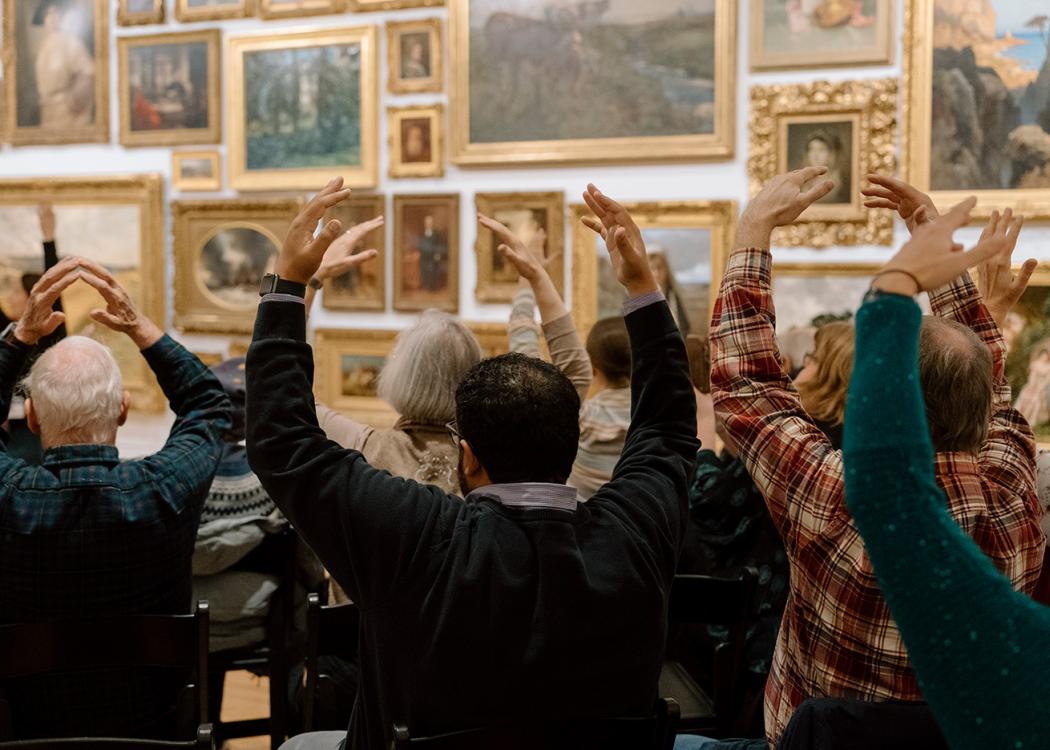 Seated people viewed from behind holding their hands above their heads in an O shape during a moment exercise in an art gallery