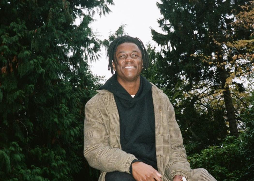 Photo of Jamal Yearwood, a young man with short black hair wearing a long tan coat, sitting on a wooden post among large evergreen trees