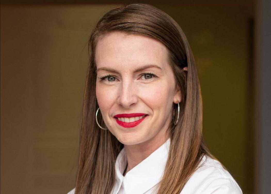 A crop of a professional headshot style photo of a smiling woman with brown hair wearing a white blouse