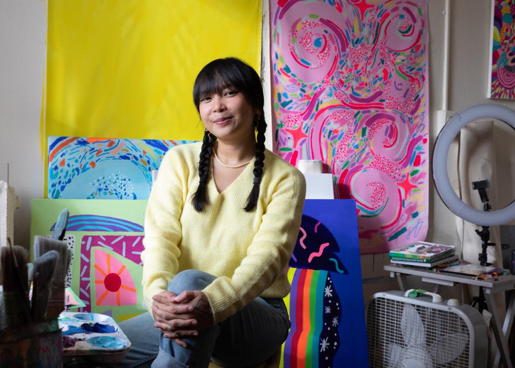 Photo of Nikita Ares, a woman with dark brown hair, smiling and sitting in her studio surrounded by her colorful paintings