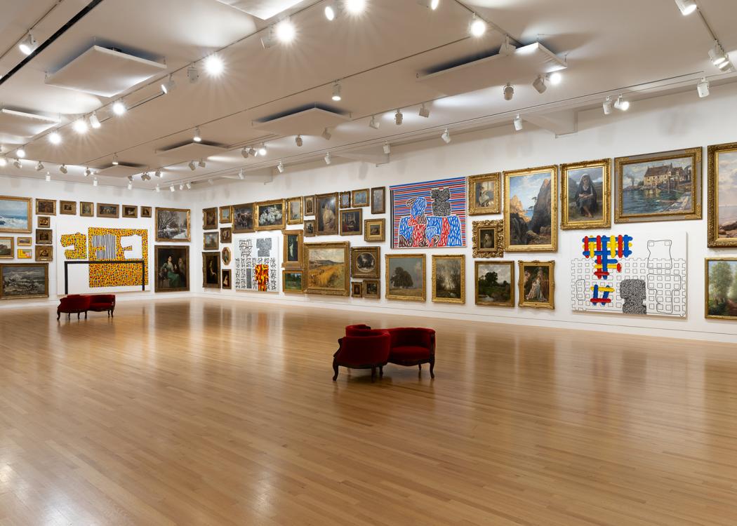 A large room with a mix of traditional and abstract paintings hung densly on the walls