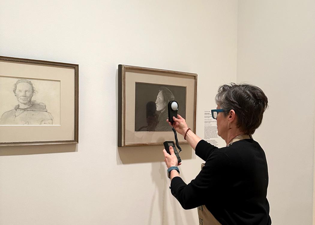 A woman holds a light meter in front of an artwork on paper hung on a gallery wall