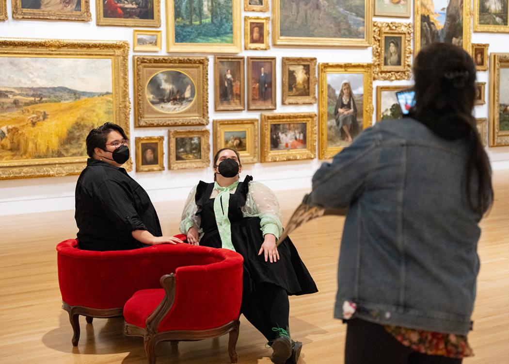 People in front of a wall covered in paintings wearing masks while sitting in a round red chair posing for a photo