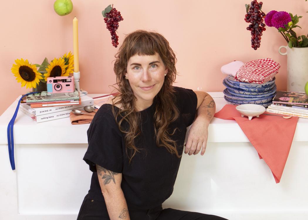 Cece Edson wearing all black while sitting on a colorful quilt with flowers, fruit, and tableware on a low shelf behind her