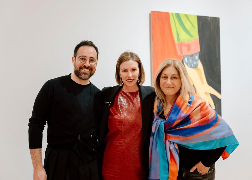 Three people standing close together and smiling in front of an artwork