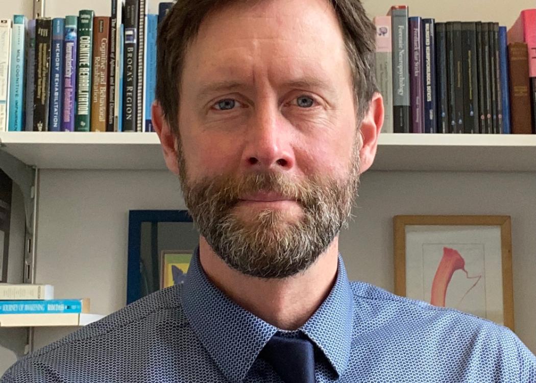 Photograph of Kris Rhoads, a man in a blue collared shirt with a blue tie, short brown hair and a beard and mustache, with a shelf of books and artwork on the wall behind him
