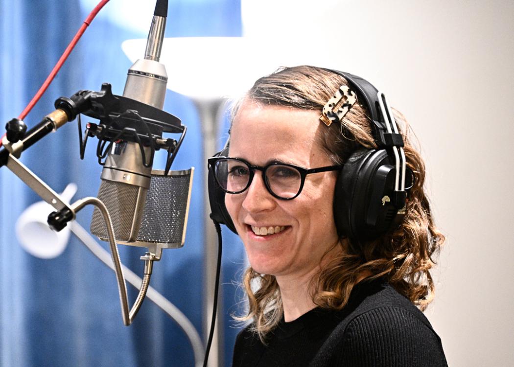 Photo of Erin Langner, a woman with shoulder length brown hair and glasses, smiling in front of a microphone in a recording studio
