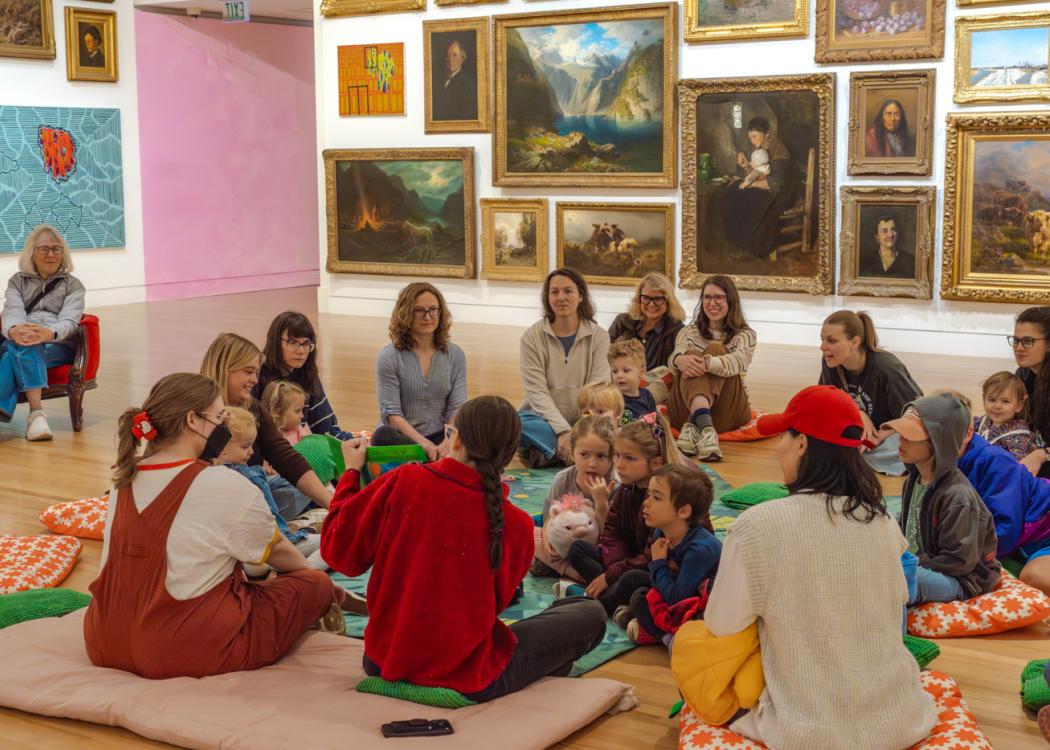 A group of adults and children sitting on the floor of a large gallery space watching someone read and show image in a picture book